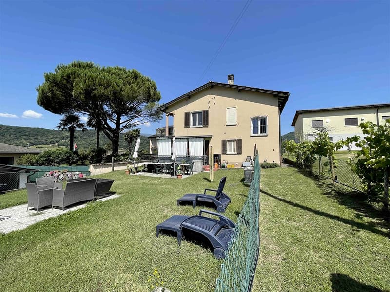 2 story house, beige exterior, large window, outdoor dining table and chairs, lounging chairs, outdoor plants, fence, expansive lawn, tree, distant building, mountains in background