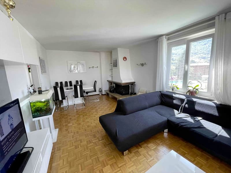 The living room has a L-shaped couch, wooden floor, white walls, and a fireplace. There is a dining area with a table and chairs. A TV with an aquarium stand and a white cabinet are in the corner. Large windows with white curtains allow natural light to f