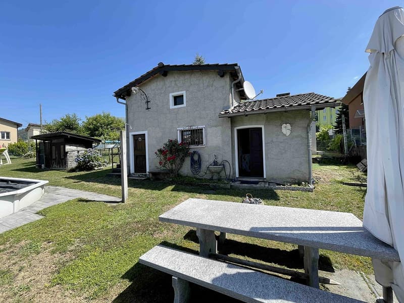 small detached house, concrete walls, clay tiled roof, two windows, door, satellite dish, garden, patio with table and bench
