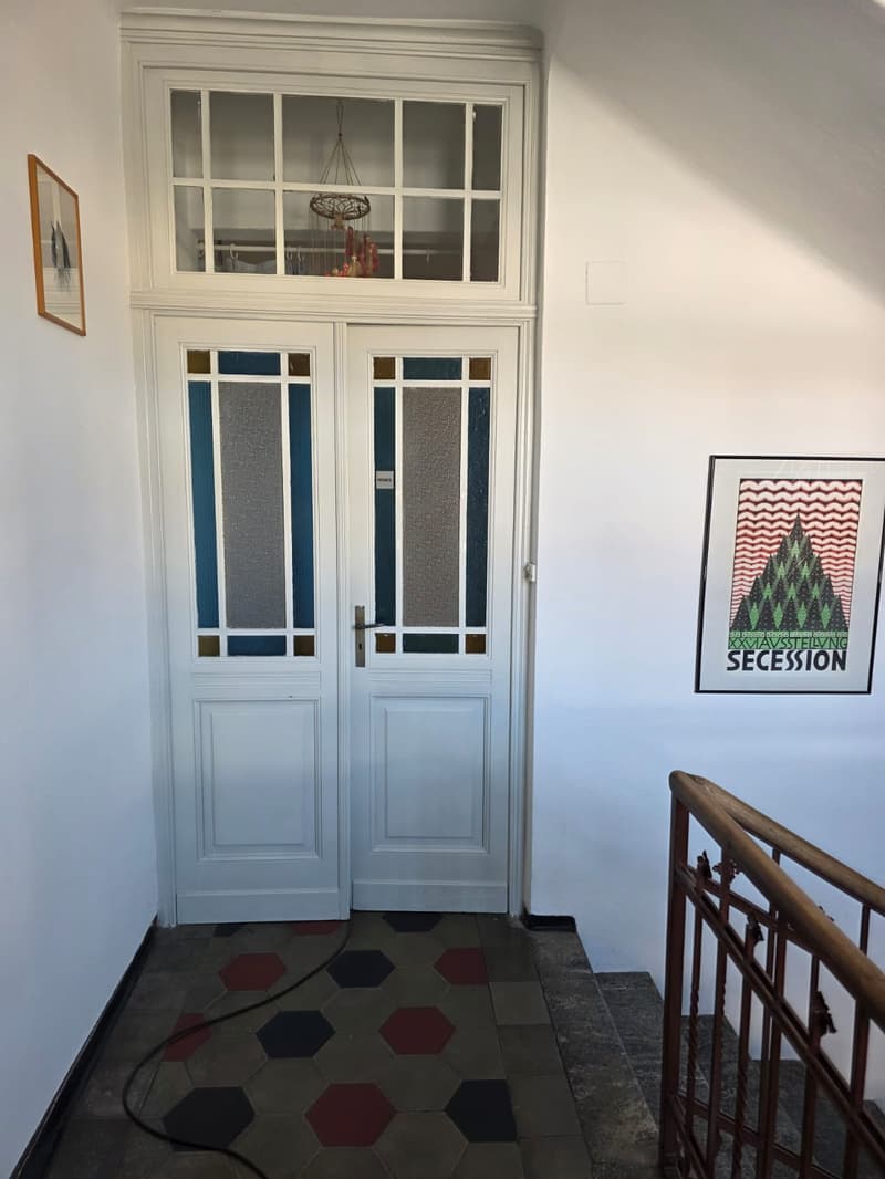 White double entrance doors with a glass window on top, geometric tiled floor, white walls, stairs leading up