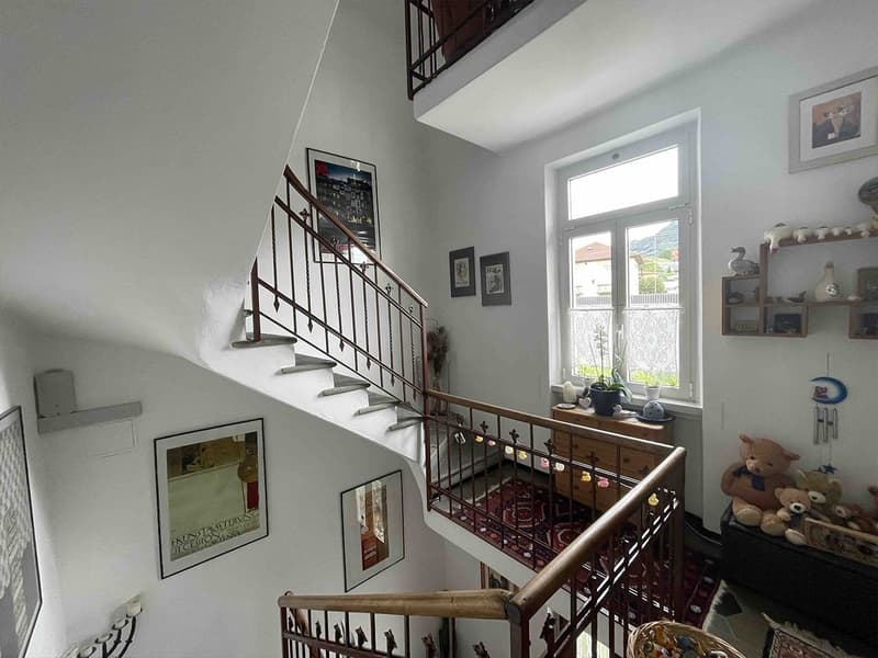 white walls, wooden stairs, metal handrails, several framed pictures on the wall, wooden cabinet, small plant on top, rug at the bottom of the stairs