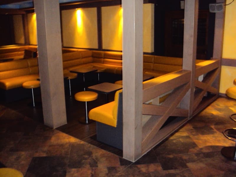 The image shows a cozy and dimly lit interior space with wooden benches and tables, as well as stools. The lighting is warm and creates a comfortable atmosphere, and there appears to be a fireplace visible in the background.