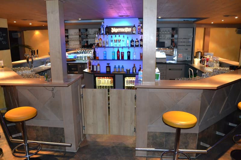 Modern bar with wooden countertops, gray cabinets, and a large selection of liquor bottles and glassware displayed on shelves. The bar has a sleek, contemporary design with yellow bar stools and a blue-lit backdrop.