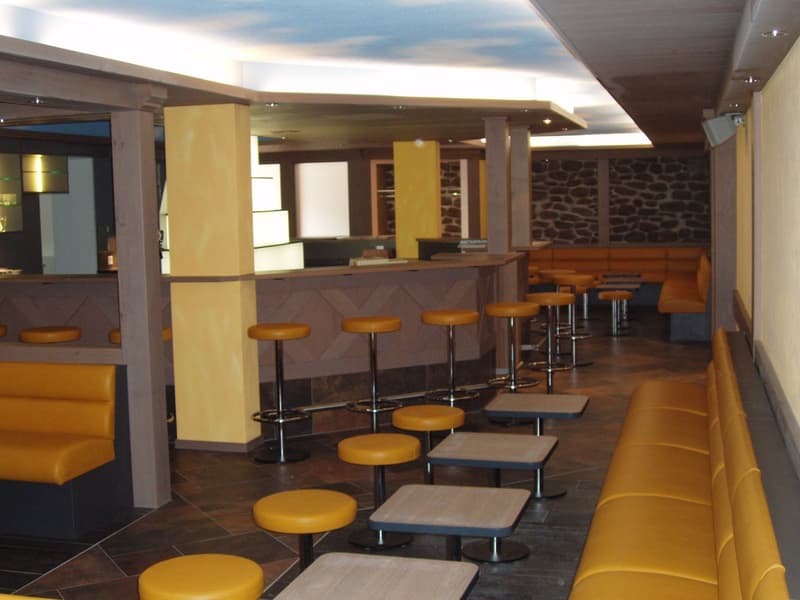 The image shows a modern and stylish bar or lounge area. The space features a long bar counter with stools, as well as several small tables and chairs for seating. The walls are made of stone, creating a cozy and rustic ambiance. The lighting is warm and