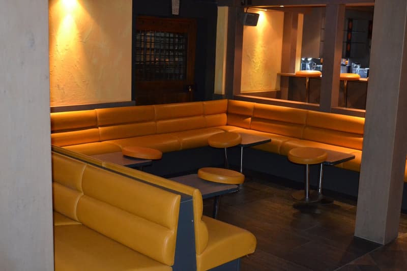 The image shows a cozy and dimly lit interior space with comfortable yellow leather seating, including a sectional sofa and individual chairs. There is a fireplace visible on the wall, adding to the warm and inviting atmosphere. The space also features wo
