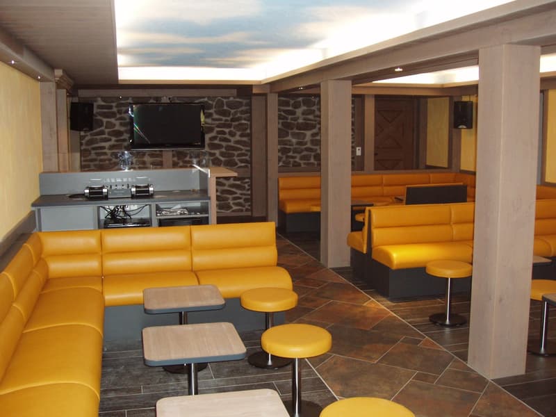 Large open-concept living space with stone walls, yellow leather sofas, wooden tables and stools, and a TV mounted on the wall. The room has a cozy and modern feel with a fireplace visible.