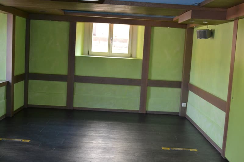 The image shows a large, open room with green walls and dark wooden beams. The room has a window and a TV mounted on the wall. The floor is covered with dark wooden planks.