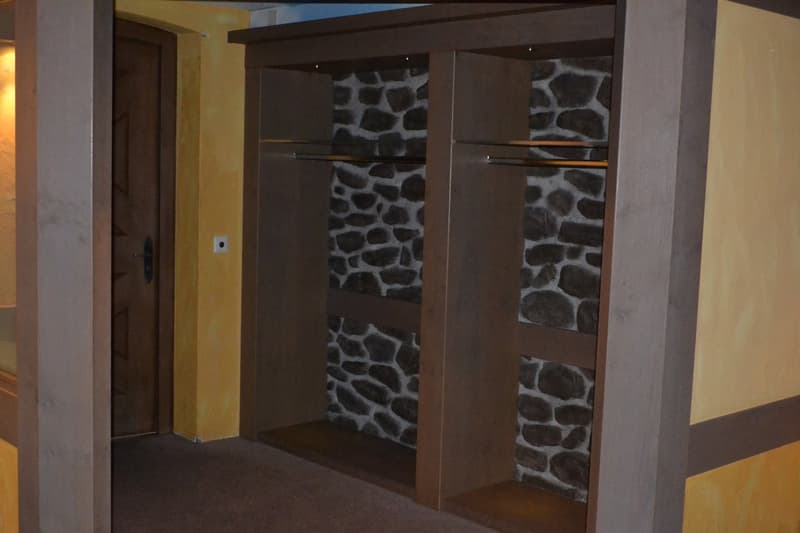 The image shows a room with a stone wall feature. The room has a wooden frame and a door leading to another area. The walls are painted in a light color, and the floor appears to be carpeted.