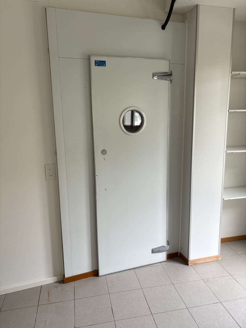 The image shows a white metal door with a circular window. The room has a tiled floor and appears to be a utility or storage room.