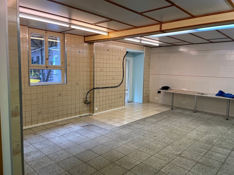 The image shows a large room with tiled floors and walls. The room has a window, a table, and some storage shelves. The ceiling has a wooden beam structure. The overall appearance suggests this could be a utility or laundry room.