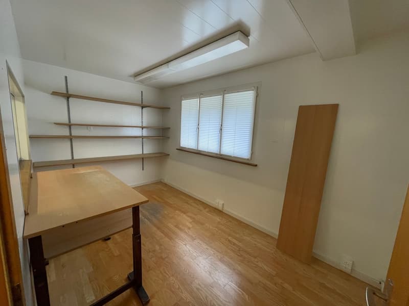 Empty room with hardwood floors, white walls, a wooden desk, shelves on the wall, a fluorescent light, and a window with white blinds.