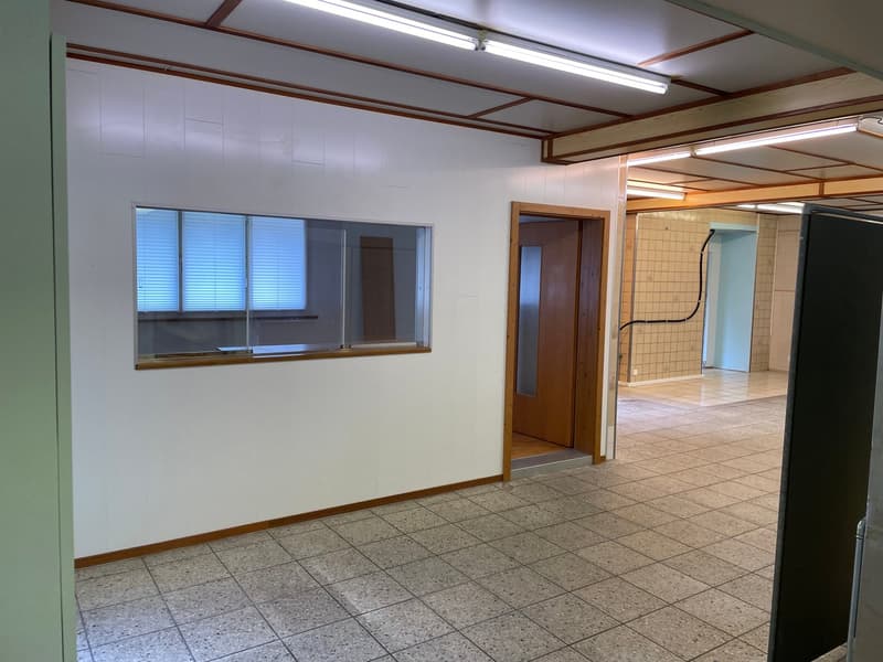 The image shows a large open office space with a tiled floor, white walls, and wooden beams on the ceiling. There is a window with blinds and a doorway leading to another room. The space appears to be a commercial or office setting.