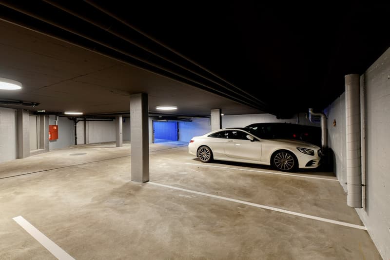 Underground parking area with concrete flooring, two parked cars, pillars, and ceiling lights