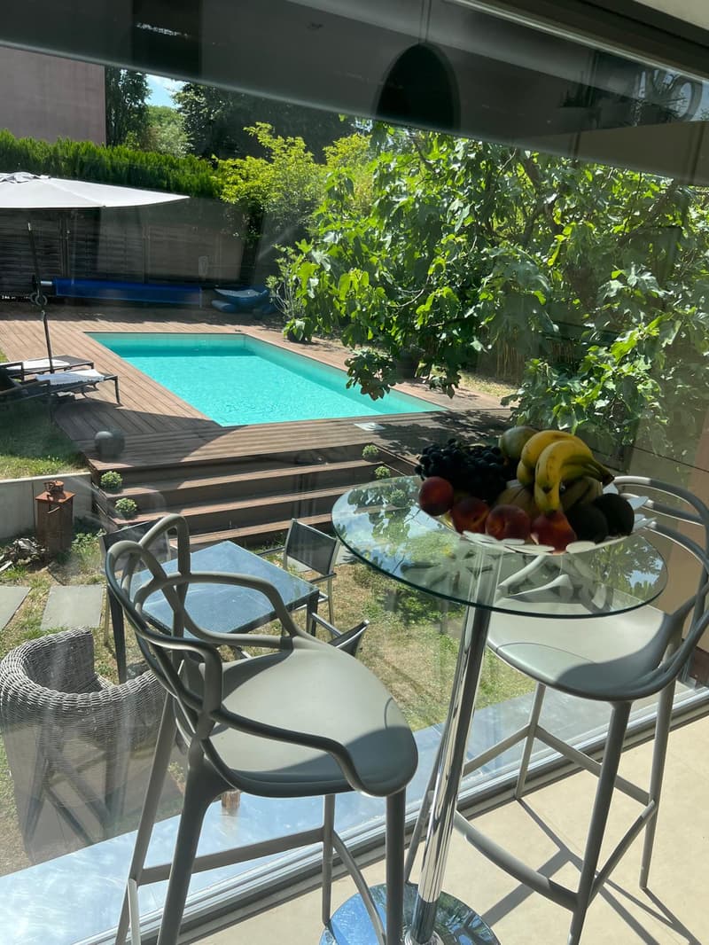 Glass table with fruit bowl, chairs, glass wall, wooden deck, swimming pool, lounge chairs, umbrella