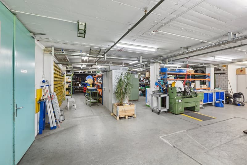 The image shows a large industrial warehouse or workshop space. The space has a concrete floor, exposed ceiling with pipes and lighting, and shelving units along the walls filled with various tools and equipment. There are also some larger machinery and e