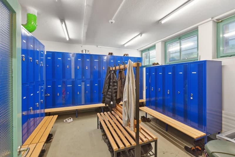 The image shows a locker room with multiple rows of blue metal lockers lining the walls. There are wooden benches and coat racks in the center of the room, where people can store their belongings and change clothes. The room appears to be well-lit and has