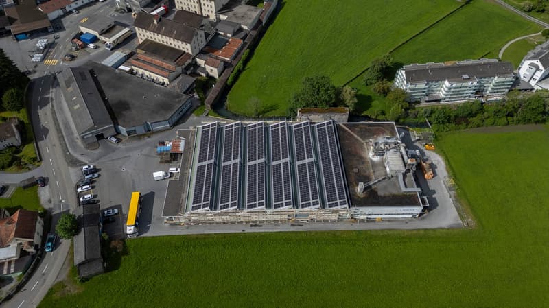 Large commercial building with a rooftop covered in solar panels, surrounded by a paved parking area and grassy fields. The building appears to be an industrial or warehouse facility located in an urban area.