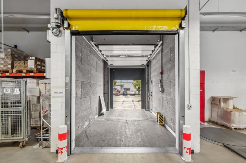 The image shows the interior of a warehouse or industrial facility. There is a large yellow roller door that appears to be the main entrance, leading to an outdoor area visible through the open doorway. The walls are made of concrete blocks, and there are