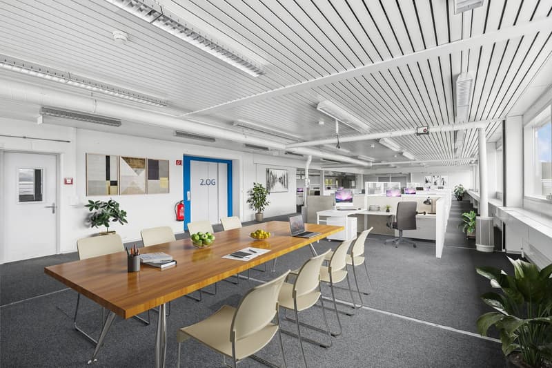 Open office space with large wooden conference table, chairs, computers, and plants. Ceiling has white paneling and lighting fixtures. Walls have artwork and signage. Appears to be a modern, well-equipped office environment.