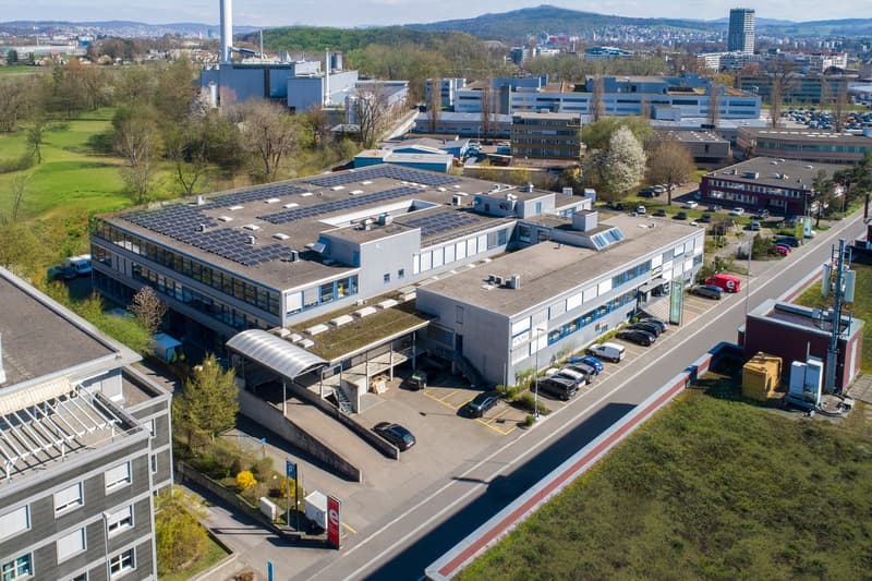 Modern building with solar panels on the roof, several parking spots on the premises, surrounded by other buildings and greenery