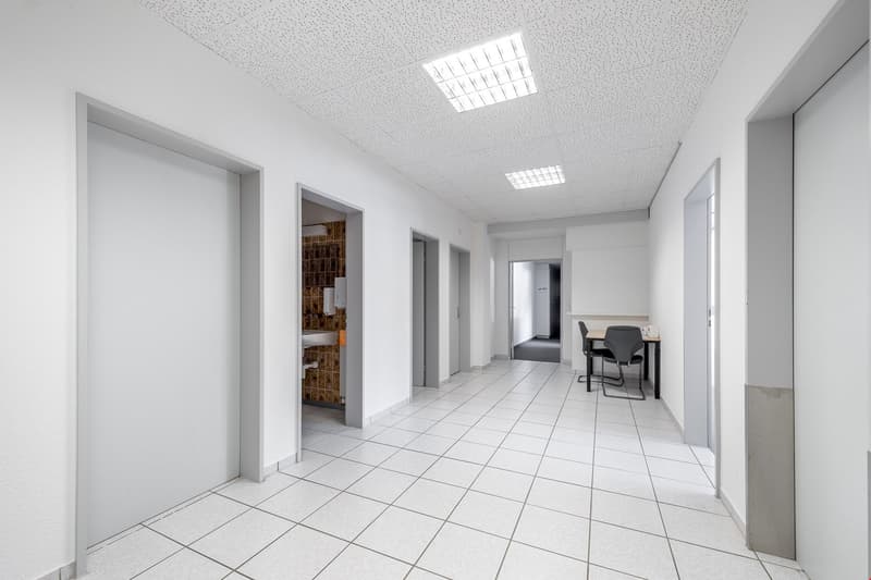 White tile flooring, white walls, grey ceiling, hallway with open doors, one room has a sink, one room has a table and chairs.