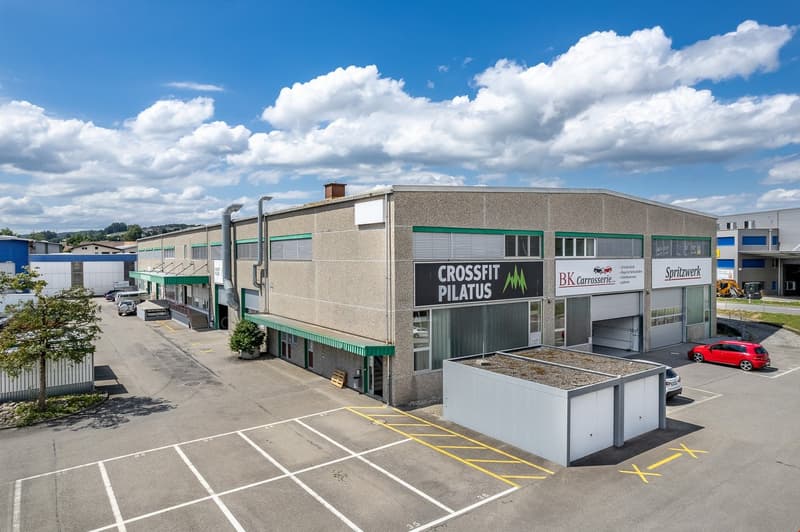 A spacious commercial building with a flat roof, multiple windows, large garage doors, signage for CROSSFIT PILATUS, several cars parked, trees and a grassy area in the foreground, and another building visible in the background.