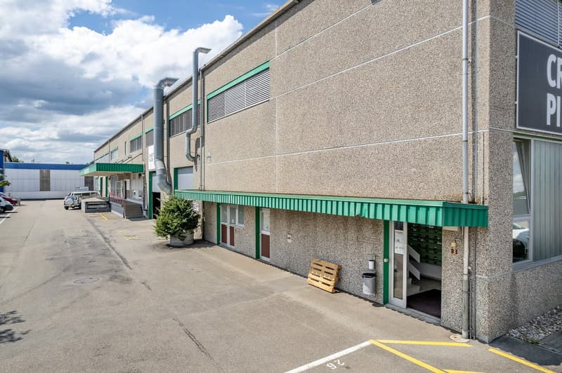 Industrial building with green awnings and an entrance, parking lot, trash bin, and plants in front.