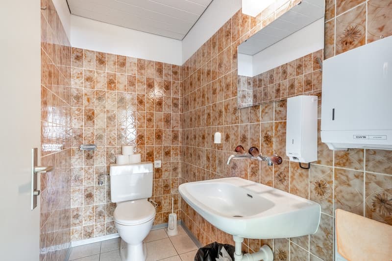 small bathroom with tile walls, white toilet, sink with mirror, toilet paper holder, hand towel holder, trash bin, toilet brush