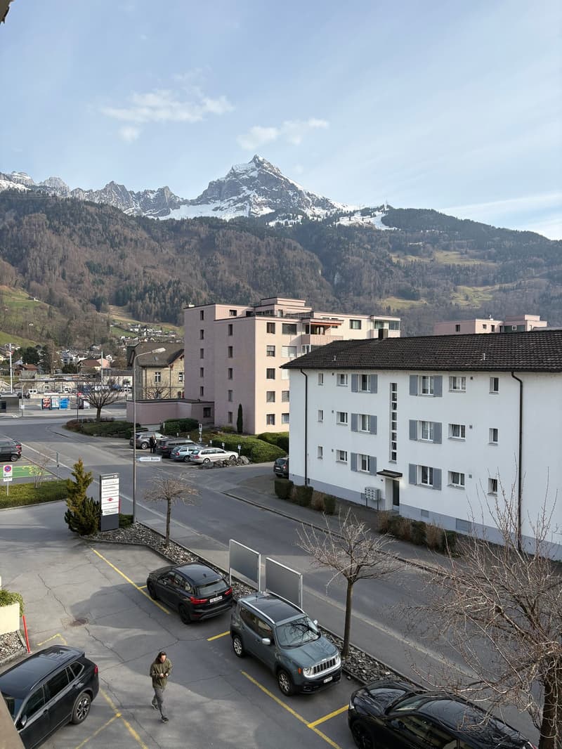 white building with several windows, pink building with several windows, many cars parked in parking lot, person walking, multiple trees, mountains with snow in background
