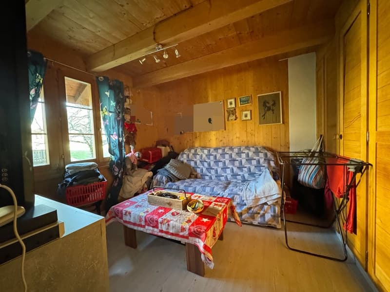 A room with wooden walls, a couch, a coffee table, a box, a basket, a clothes rack, a lamp, picture frames, a window, and curtains.