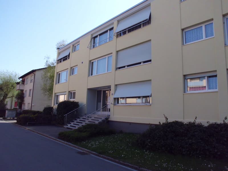 A yellow building with multiple windows and a staircase leading to the entrance. The building has a flat roof and is surrounded by bushes and grass.