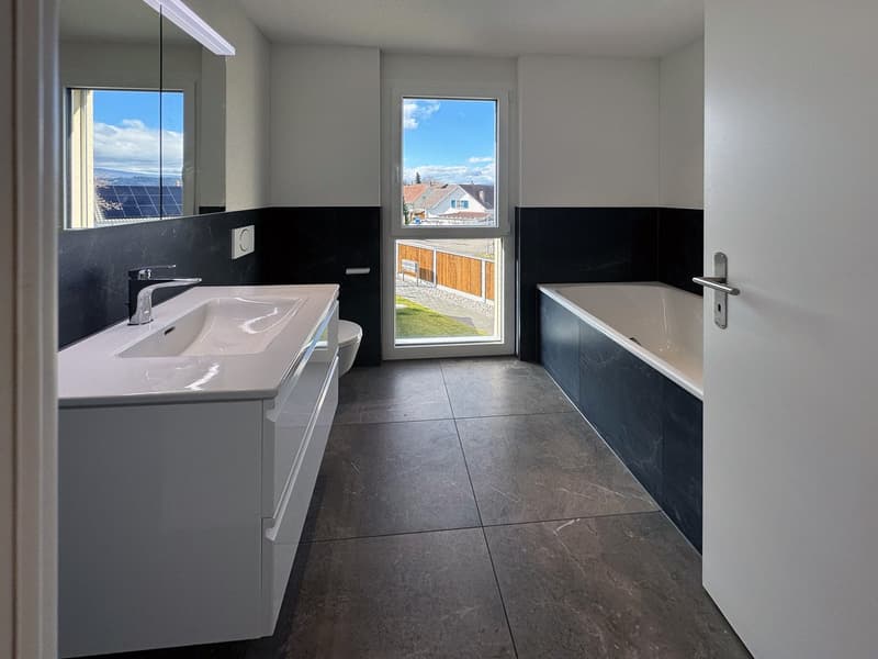 Large white modern bathroom with glass wall and black tiles, large window with view, white vanity with sink, and bathtub