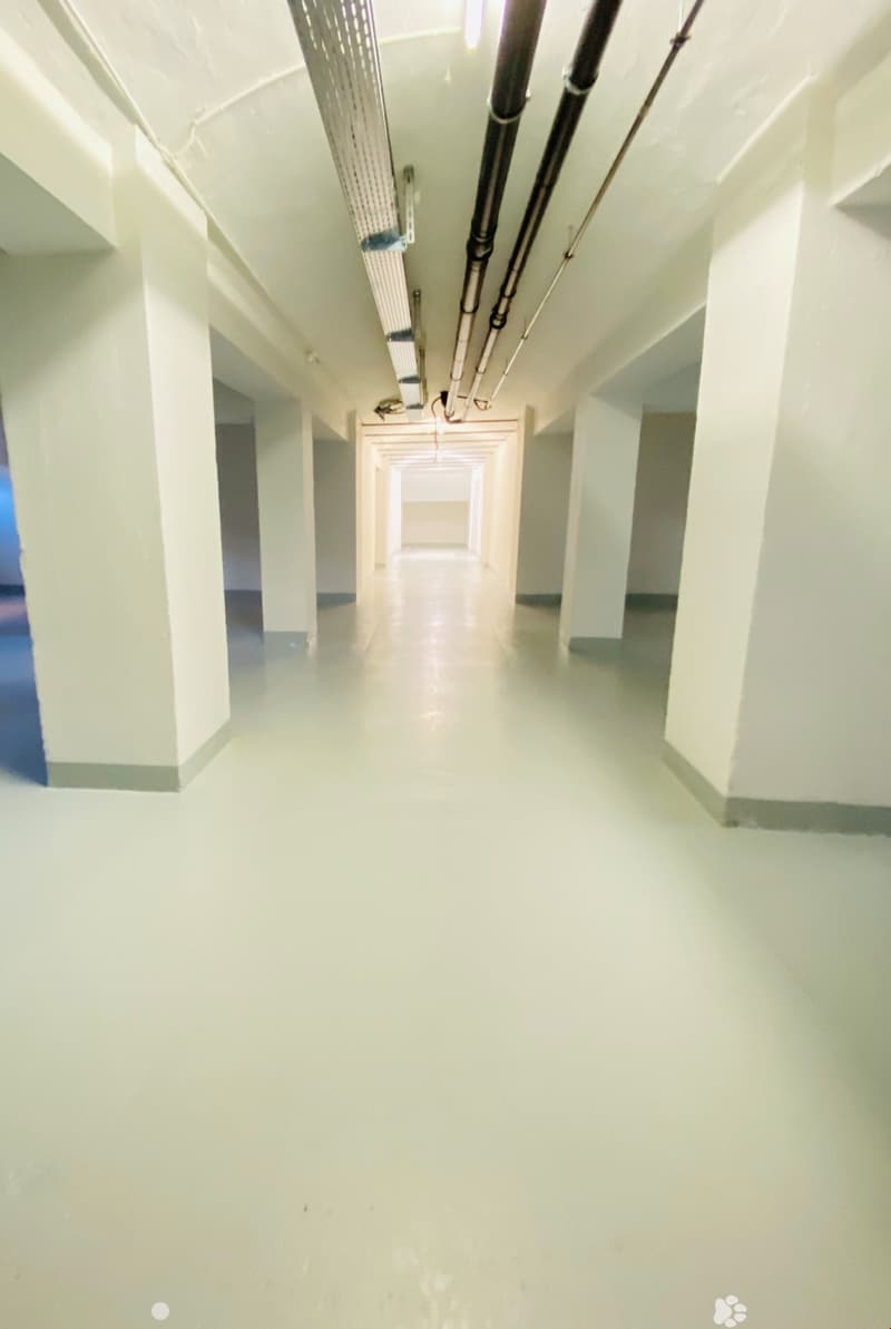 Long, narrow hallway with white walls and floors, recessed lighting, and pipes running along the ceiling.