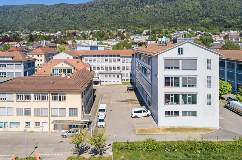 The image shows an aerial view of a city or town, with a mix of residential and commercial buildings. The buildings have a variety of architectural styles, with some older structures and some more modern ones. There are trees and greenery visible througho