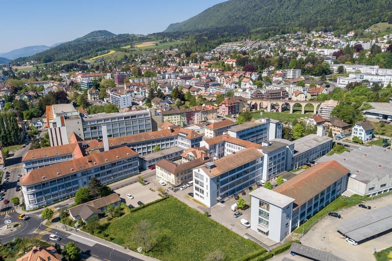 Aerial view of a city nestled in a valley surrounded by mountains. The city has a mix of modern and traditional buildings, with red-tiled roofs and multi-story structures. There are roads, parking lots, and green spaces visible. The landscape features for