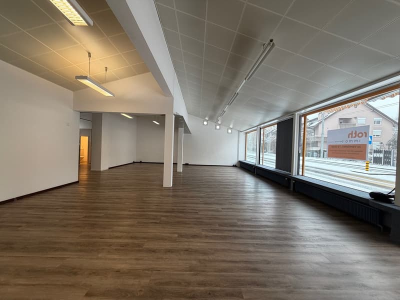 An empty room with hardwood floors, white walls, and fluorescent ceiling lights, large windows on one side. A signboard is positioned in front of the windows.