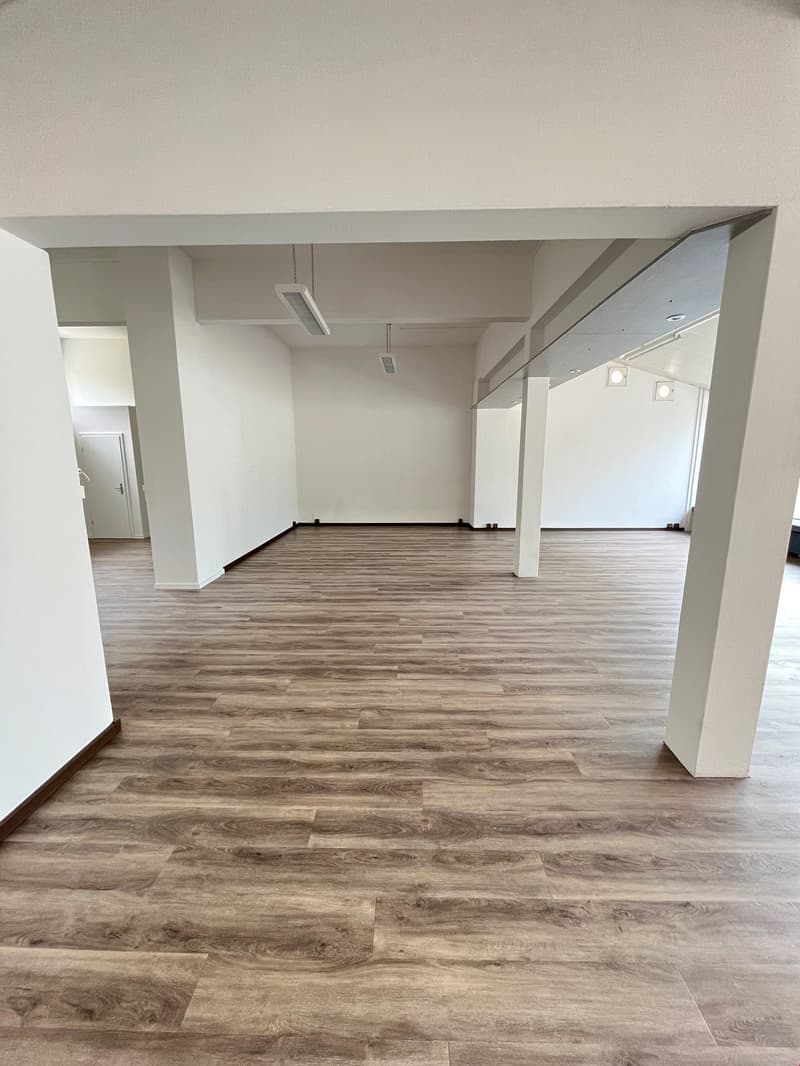 The image shows a large, open office space with a high ceiling and white walls. The floor is covered in a wood-like laminate flooring. The space appears to be empty and ready for use, with track lighting installed overhead.