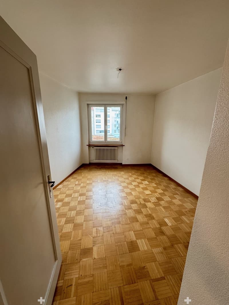 The image shows a spacious bedroom with a large window, hardwood parquet flooring, and plain white walls. The room appears to be empty, with no furniture or decor visible.