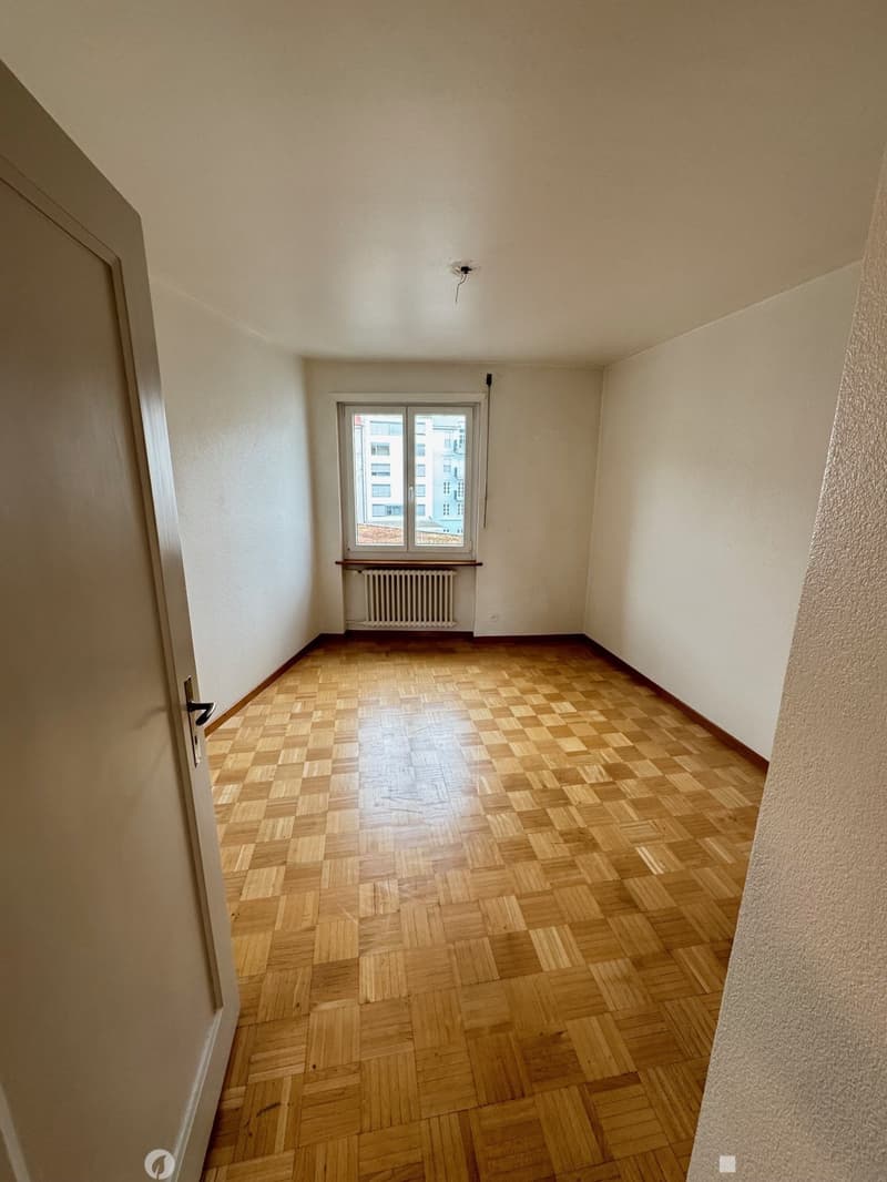 The image shows a spacious bedroom with a large window, hardwood parquet flooring, and plain white walls. The room appears to be empty, with no furniture or decor visible.