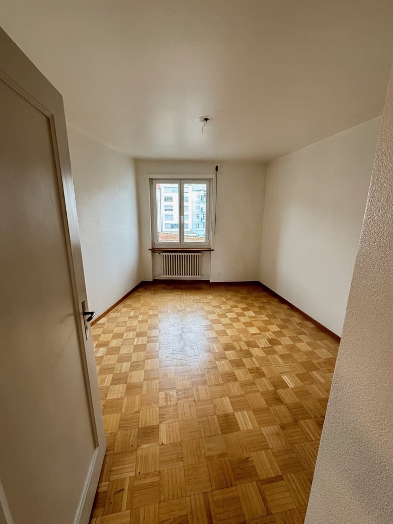 The image shows a spacious bedroom with a large window, hardwood parquet flooring, and plain white walls. The room appears to be empty, with no furniture or decor visible.