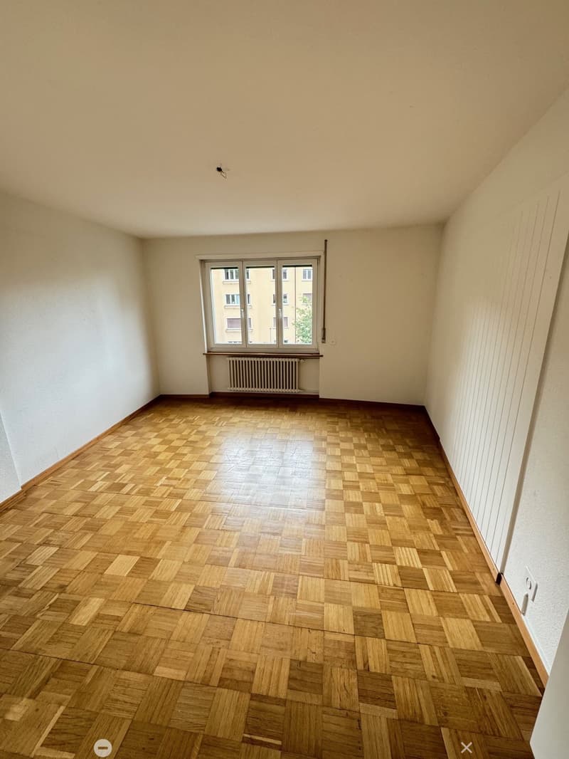 Spacious bedroom with hardwood parquet flooring, large window, and radiator.