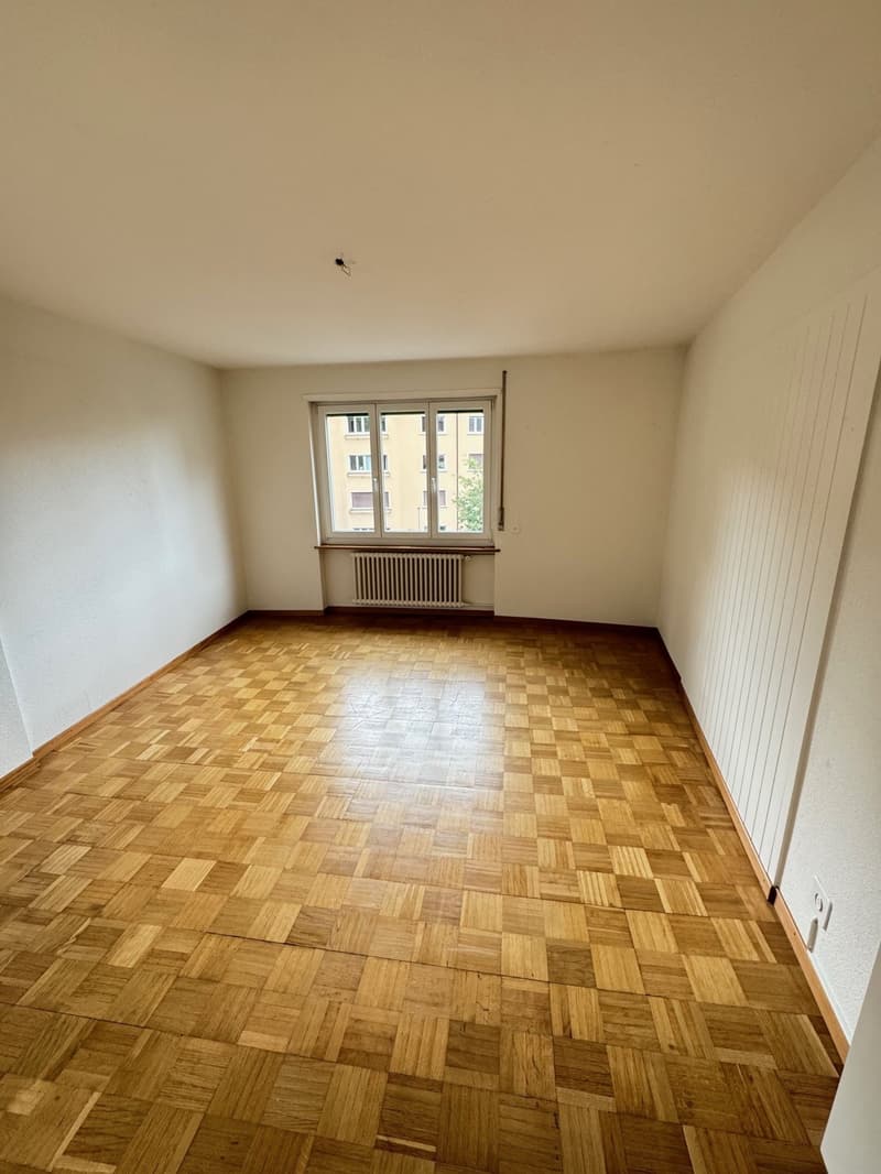 Spacious bedroom with hardwood parquet flooring, large window, and radiator.