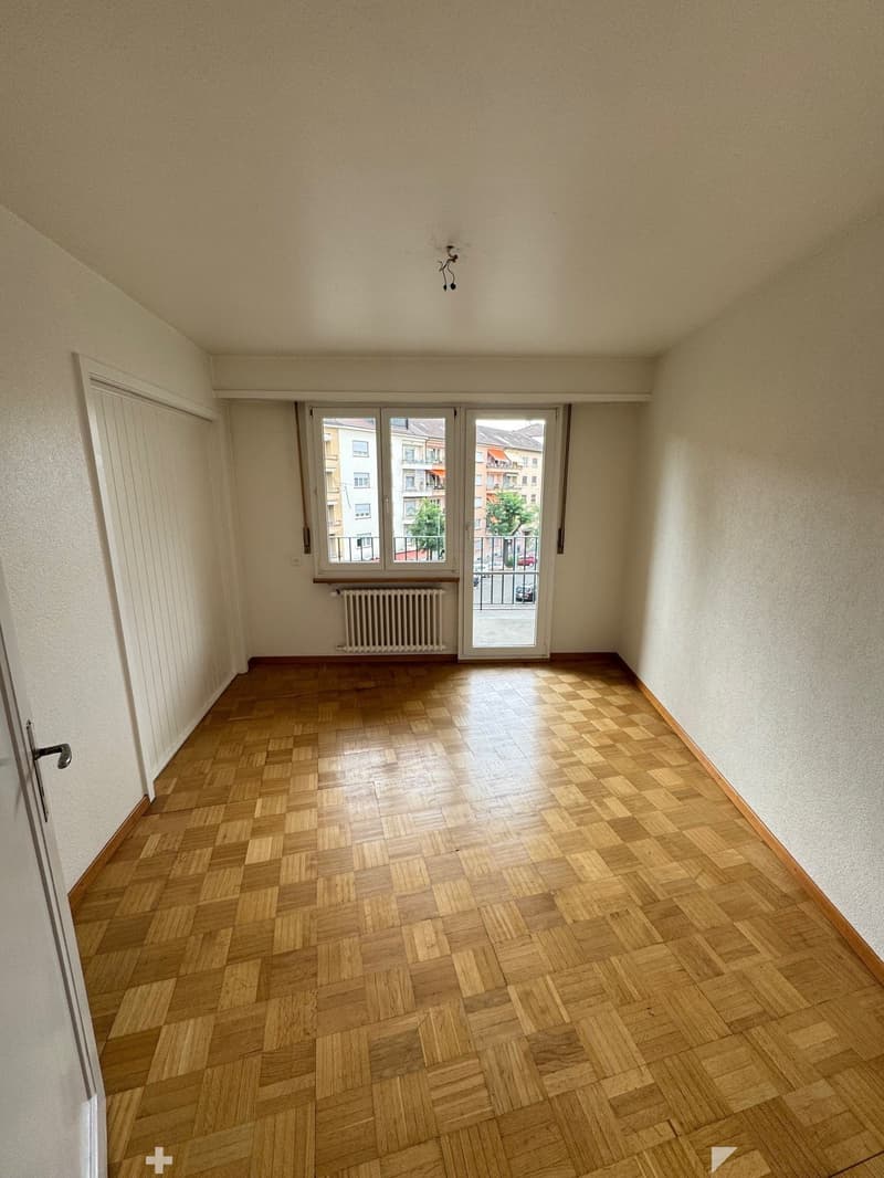 This image shows an empty room with a hardwood parquet floor, a sliding glass door leading to a balcony or terrace, and a radiator on the wall. The room has plain white walls and a simple light fixture on the ceiling.