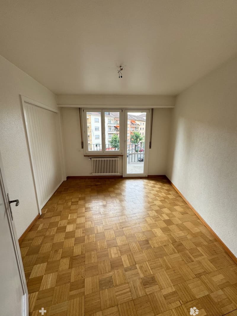 This image shows an empty room with a hardwood parquet floor, a sliding glass door leading to a balcony or terrace, and a radiator on the wall. The room has plain white walls and a simple light fixture on the ceiling.
