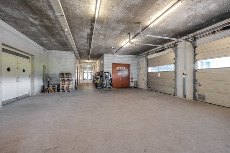 The image shows a large, open commercial space with concrete walls and ceiling, exposed pipes and lighting fixtures, and a garage door. The space appears to be used for storage or a workshop, with pallets and equipment visible. There is a door leading to