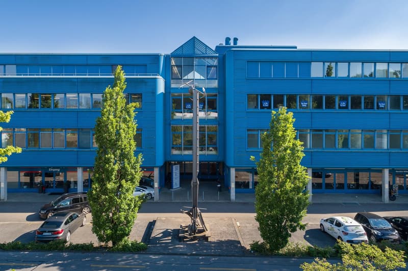 A large blue commercial building with many windows, surrounded by trees and parked cars in the foreground.