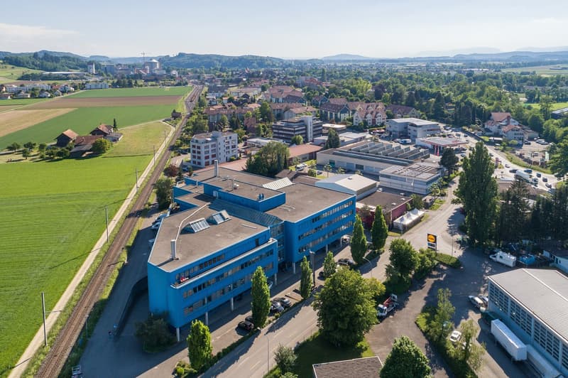 Aerial view of a city with a mix of residential and commercial buildings, surrounded by green fields and hills. The city has a train station and various commercial and industrial buildings, including a large blue office building with a parking lot in the