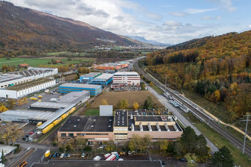 Industrial complex with multiple buildings, parking lots, road, railways, and surrounded by trees and mountains