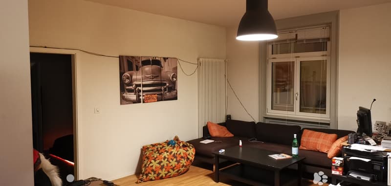 sleek lamp, modern black couch, patterned ottoman, rustic wood floor, picture of car on wall, white radiator, window with white blinds