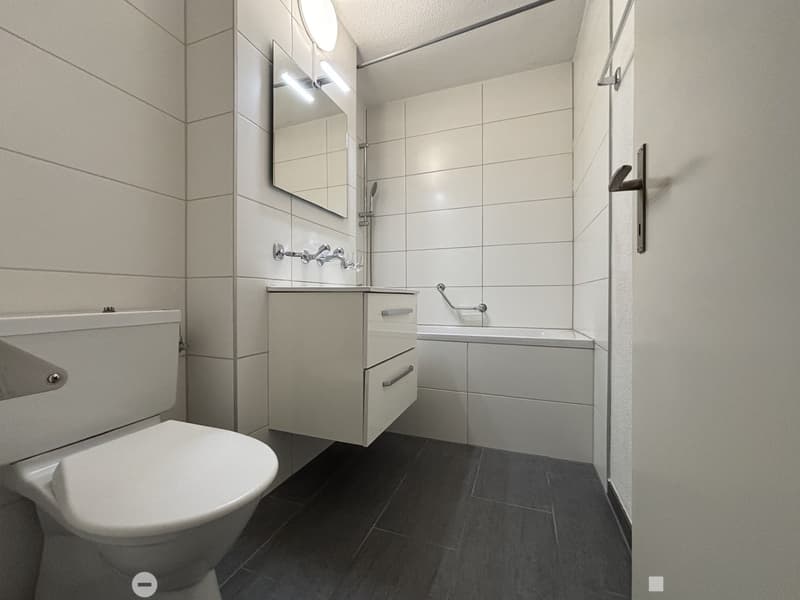 Modern bathroom, white tiled walls, white sink, white toilet, white bathtub, white shelves, bathtub grab bar, illuminated mirror
